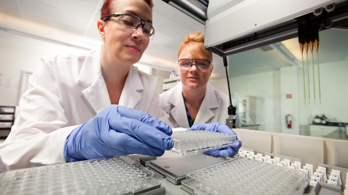 A faculty member and grad student in lab coats and safety goggles work with lab equipment.